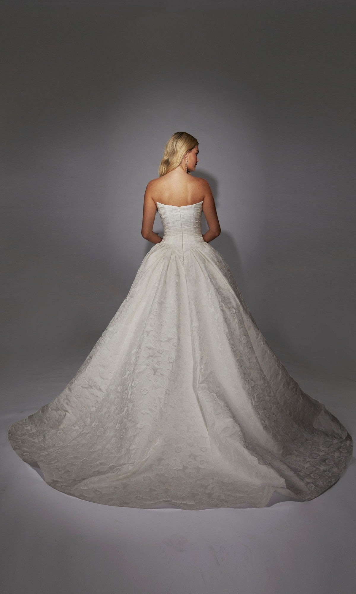 A woman stands facing away in the Alyce 7164 long white dress, a strapless gown with a full skirt and train, crafted from floral jacquard fabric, set against a neutral background.